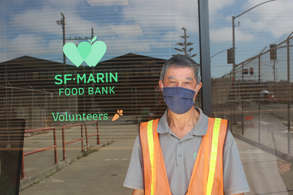 A gray haired man in a blue mask stands looking at the camera in front of a window that reads "SF Marin Food Bank Volunteers" in green lettering.